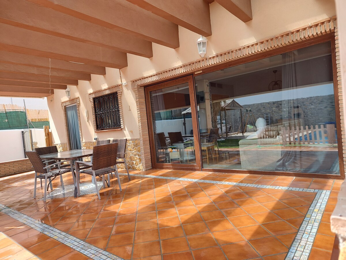 A spacious porch area is highlighted by terracotta tiles and wooden beams. A dining table with wicker chairs is positioned invitingly, providing a view of the landscaped garden and pool area beyond the large glass sliding doors.