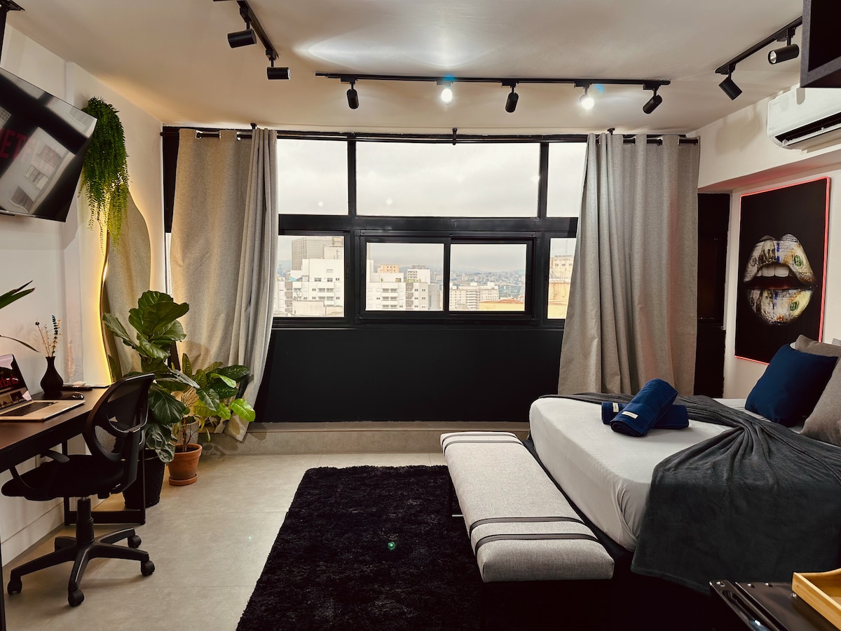 A modern loft space features a spacious bed with soft linens, complemented by a comfortable chair and desk area. Large windows offer a city view, while track lighting highlights the room's design. Greenery is present near the windows, enhancing the ambiance.