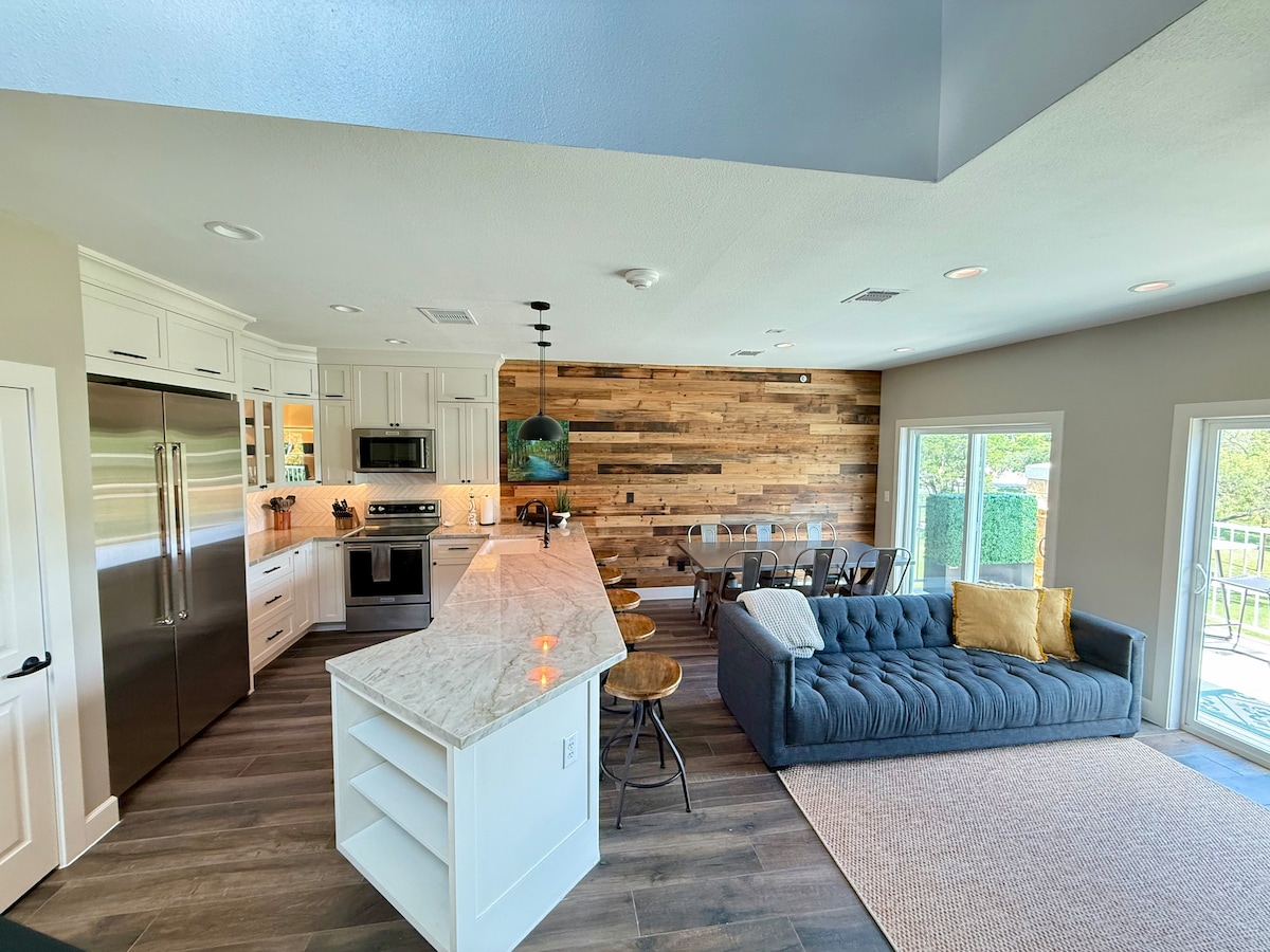 The open-concept kitchen and living area feature modern appliances and a large island with seating. A cozy sectional sofa is positioned near large windows, allowing natural light to fill the space. A dining area with a wooden table is visible in the background against a wood accent wall.