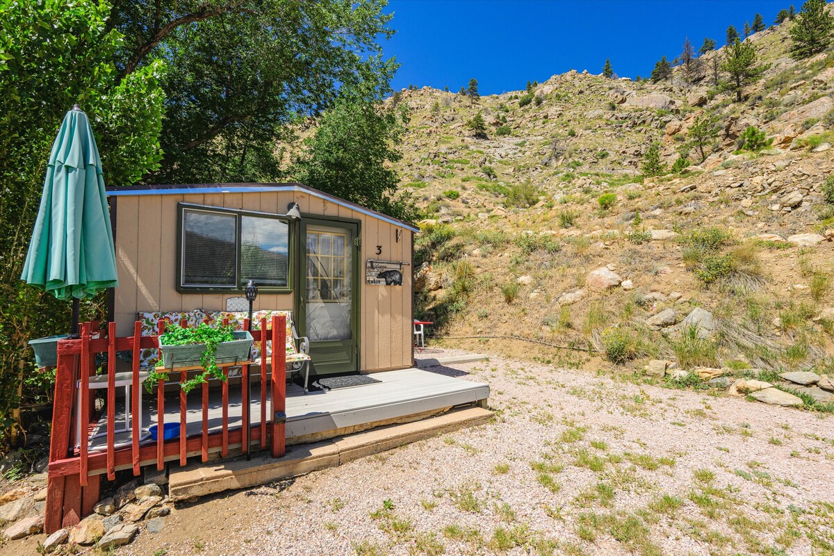 A wildlife themed cabin is set against a rocky hillside, showcasing natural landscaping with sparse vegetation. A welcoming porch supports a small table and chairs, complemented by a green umbrella. Mountain terrain rises in the background under a clear blue sky.