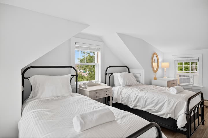 Svijetla i udobna spavaća soba na drugom katu s tri odvojena kreveta, savršena za djecu ili prijatelje. Klasični nakošeni stropovi u stilu Cape Coda, noćni ormarić u retro stilu i meka bijela posteljina. Čisto, udobno i puno prirodnog svjetla.