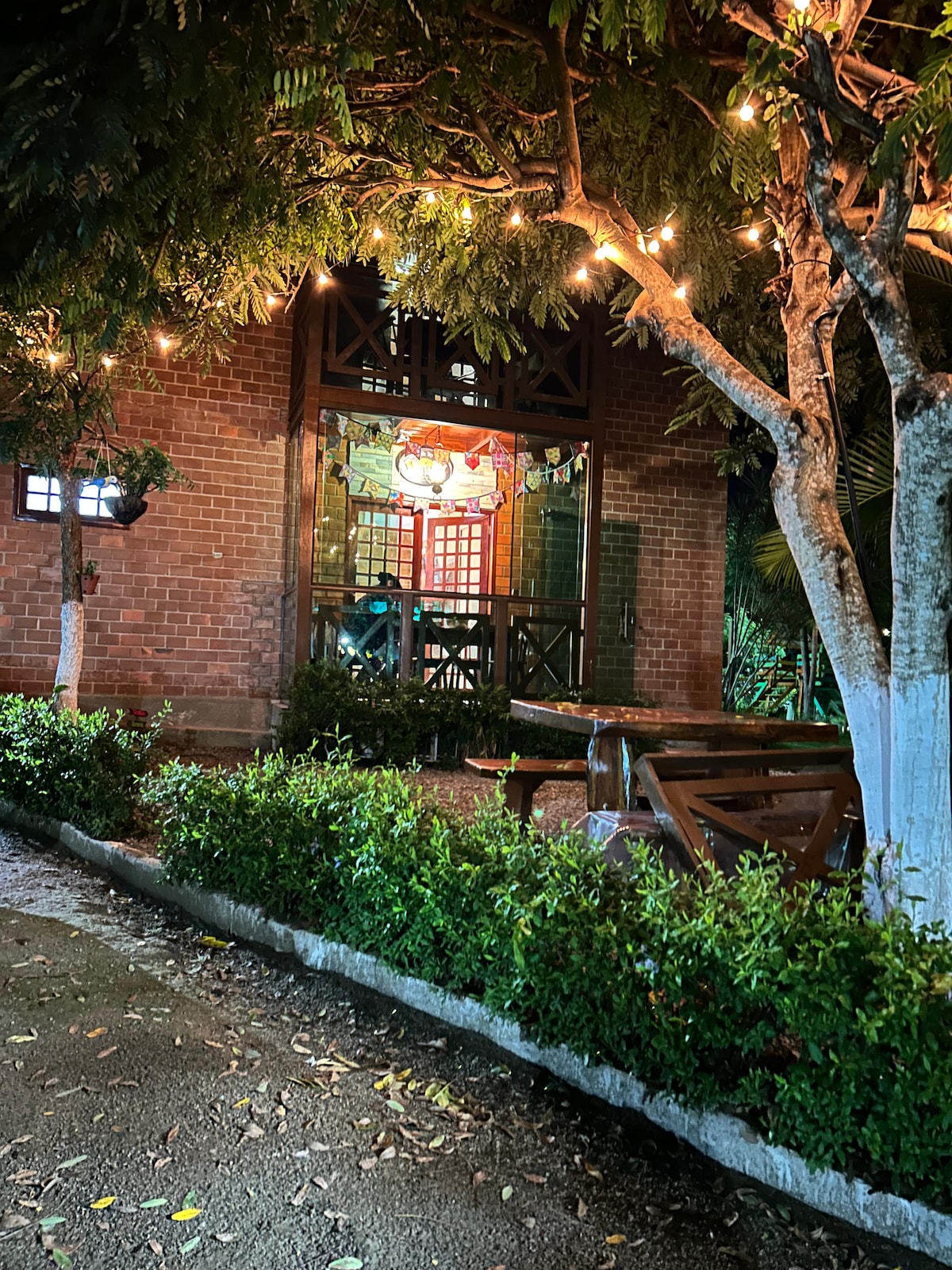 An inviting outdoor area is shown at night, featuring a wooden structure illuminated by soft lighting. Surrounding greenery adds to the serene atmosphere. In the foreground, a rustic picnic table sits beneath trees adorned with warm string lights, enhancing the inviting outdoor space.