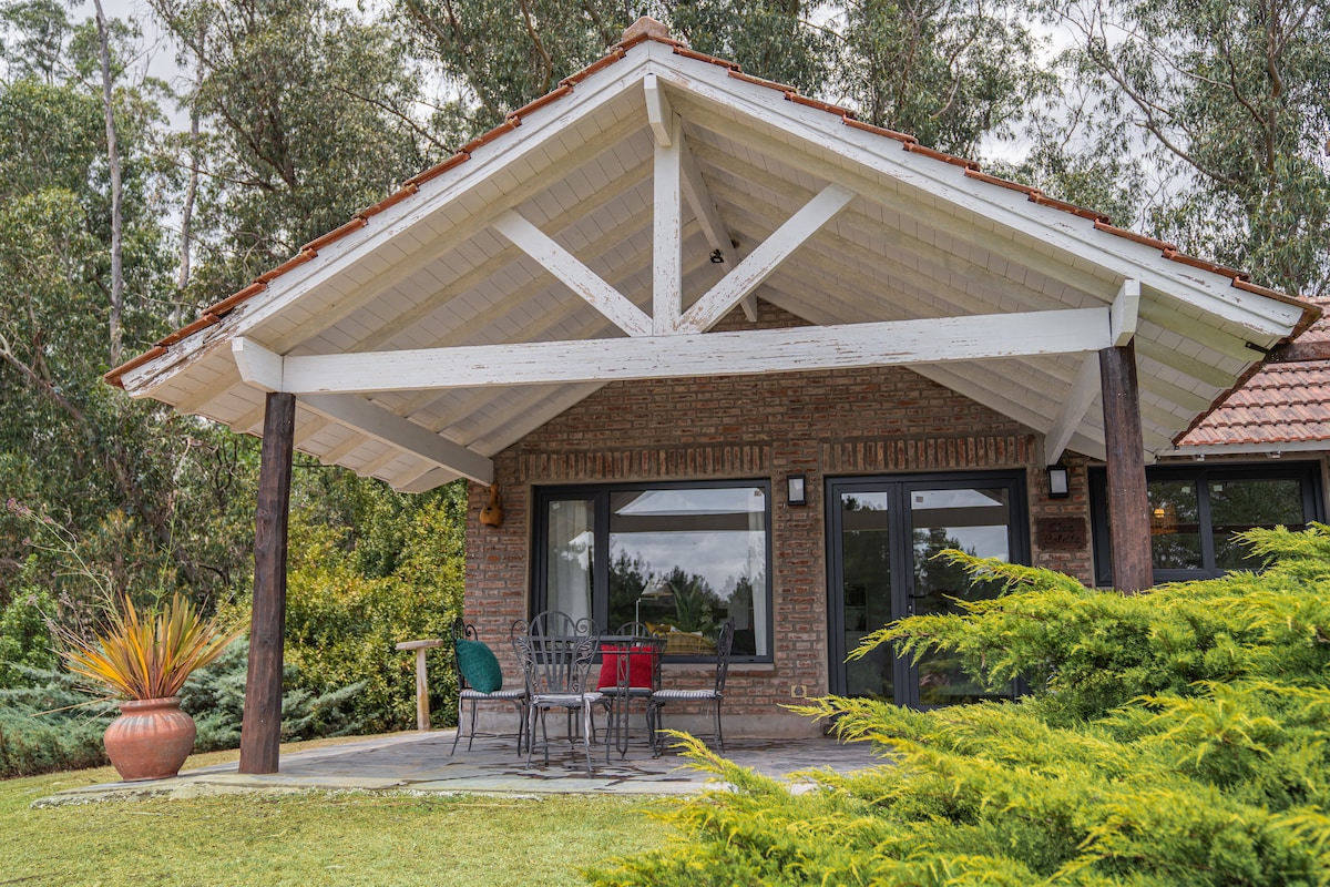 A charming outdoor seating area features a covered porch with a slanted roof supported by wooden beams. Gray metal chairs are arranged around a small table, with potted plants and greenery enhancing the natural surroundings.