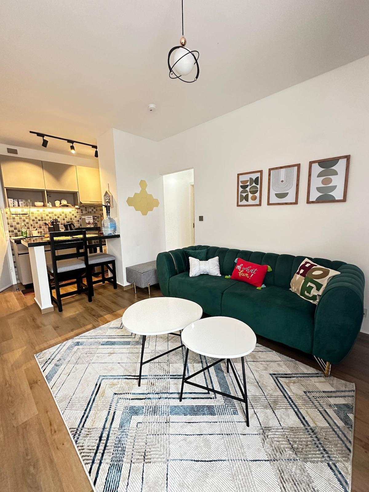A cozy living area features a green sofa adorned with decorative pillows. Two elegant side tables are positioned over a patterned rug. In the background, a kitchenette with modern cabinetry and appliances is visible. Warm light provides a welcoming ambiance.