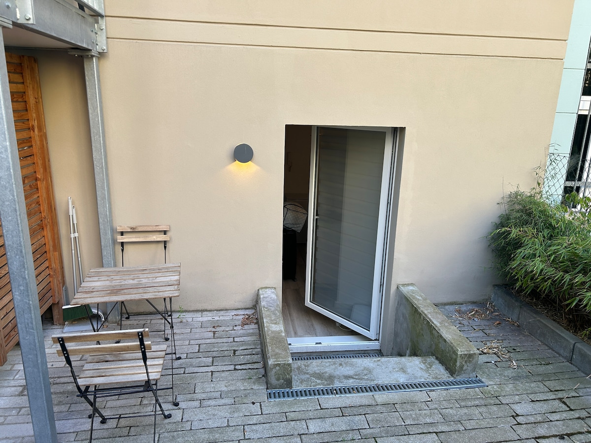 A private terrace is shown featuring a small wooden table and two chairs, positioned on stone flooring. A doorway leads inside, illuminated by a wall-mounted light. Lush greenery is visible nearby, creating a calm outdoor setting.