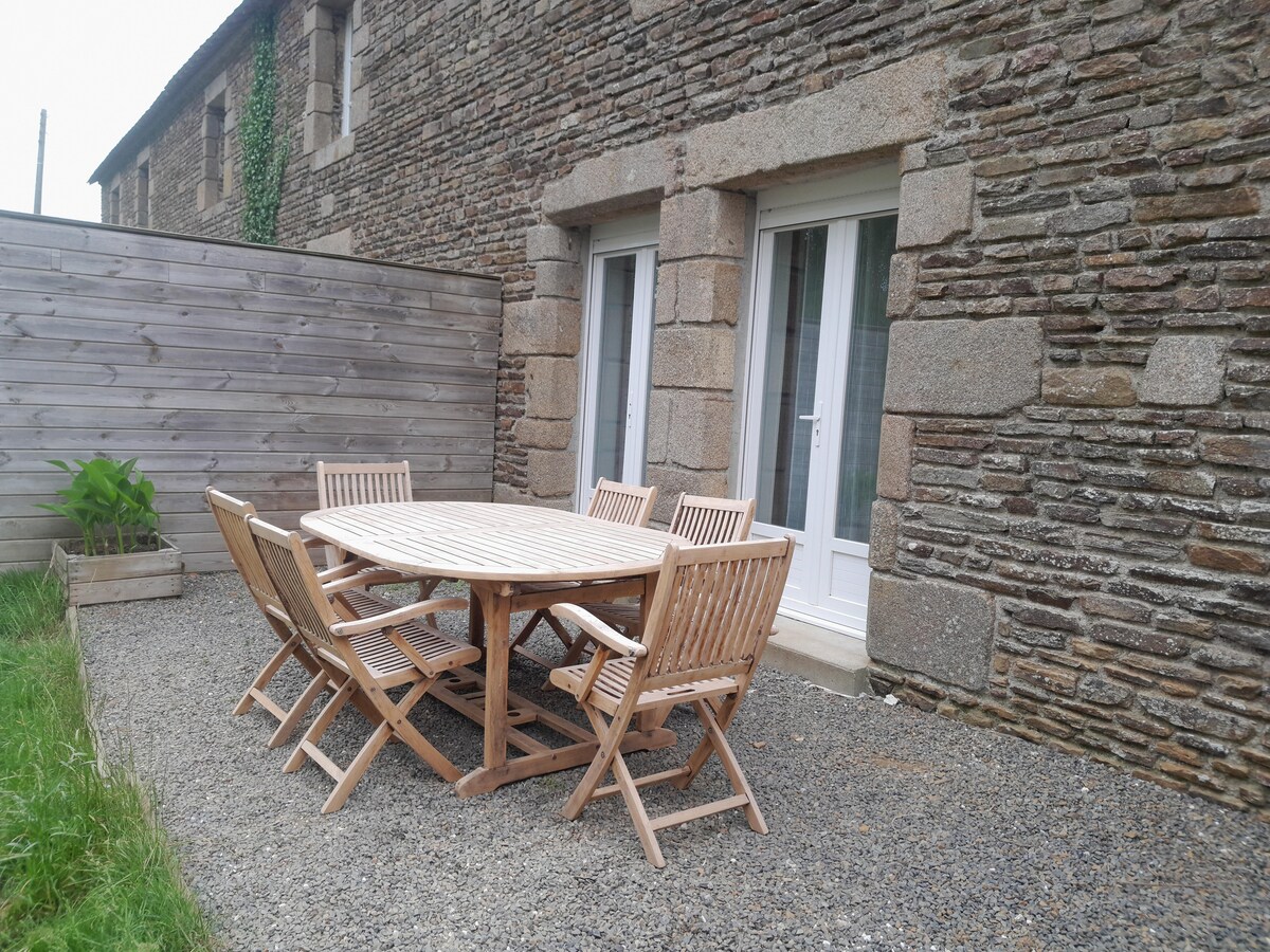 An outdoor patio area features a round wooden table accompanied by six matching chairs. The setting is bordered by a low wooden fence and a stone wall, with a small planter that adds a touch of greenery.