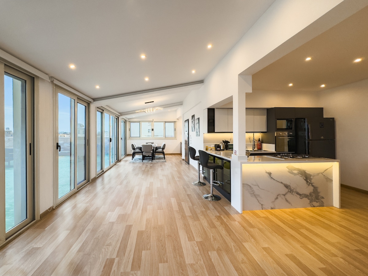 The open floor plan showcases a bright living area with large glass sliding doors, providing a view of the surrounding area. A sleek dining space is visible adjacent to a modern kitchen featuring high-end appliances. Natural light floods the space, accentuating the light wooden flooring.