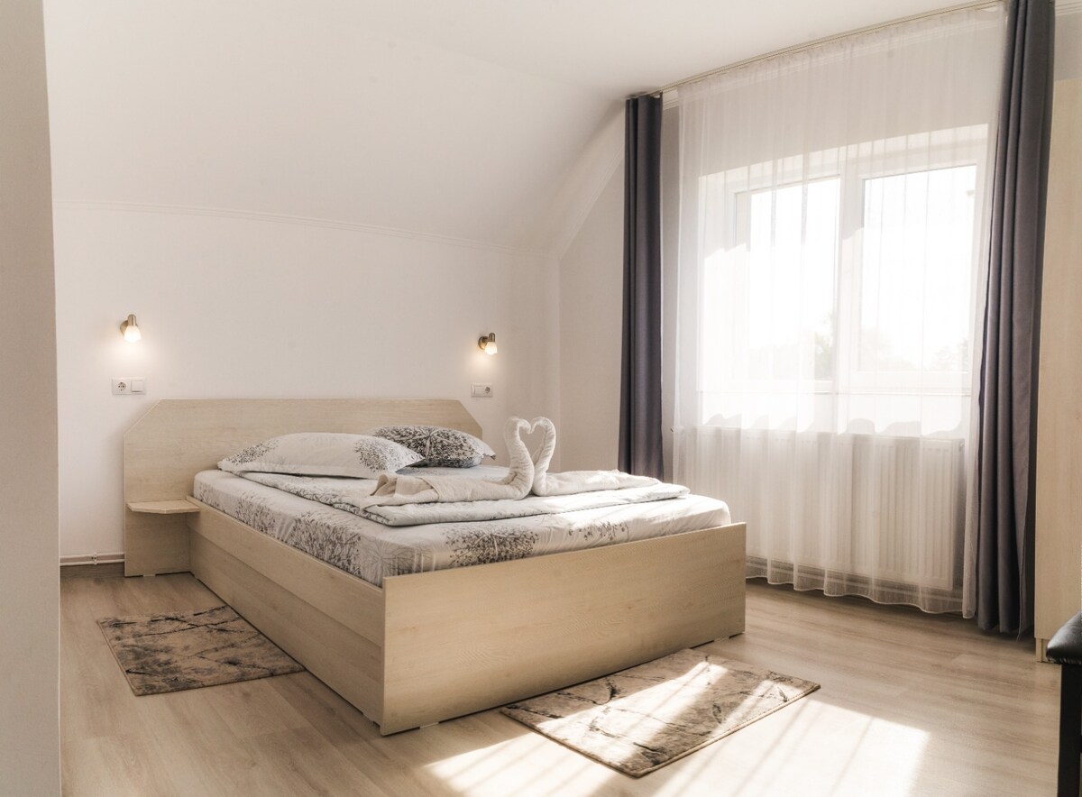 A bright and airy bedroom is presented with a spacious bed layered in soft bedding. Natural light filters through large windows, enhancing the clean and minimalist decor. Two elegant wall sconces provide gentle illumination, and the floor is adorned with simple area rugs.
