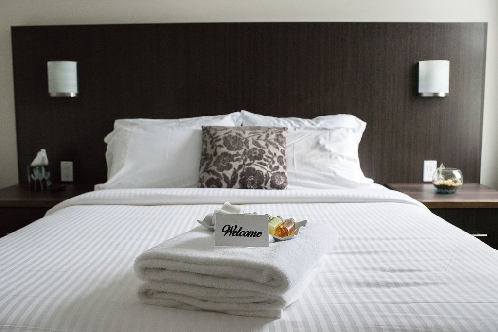 A spacious bed is adorned with crisp white linens and a decorative pillow featuring a floral pattern. Fresh towels are neatly arranged at the foot of the bed, accompanied by a welcome card and small complimentary items on a dark wood nightstand.