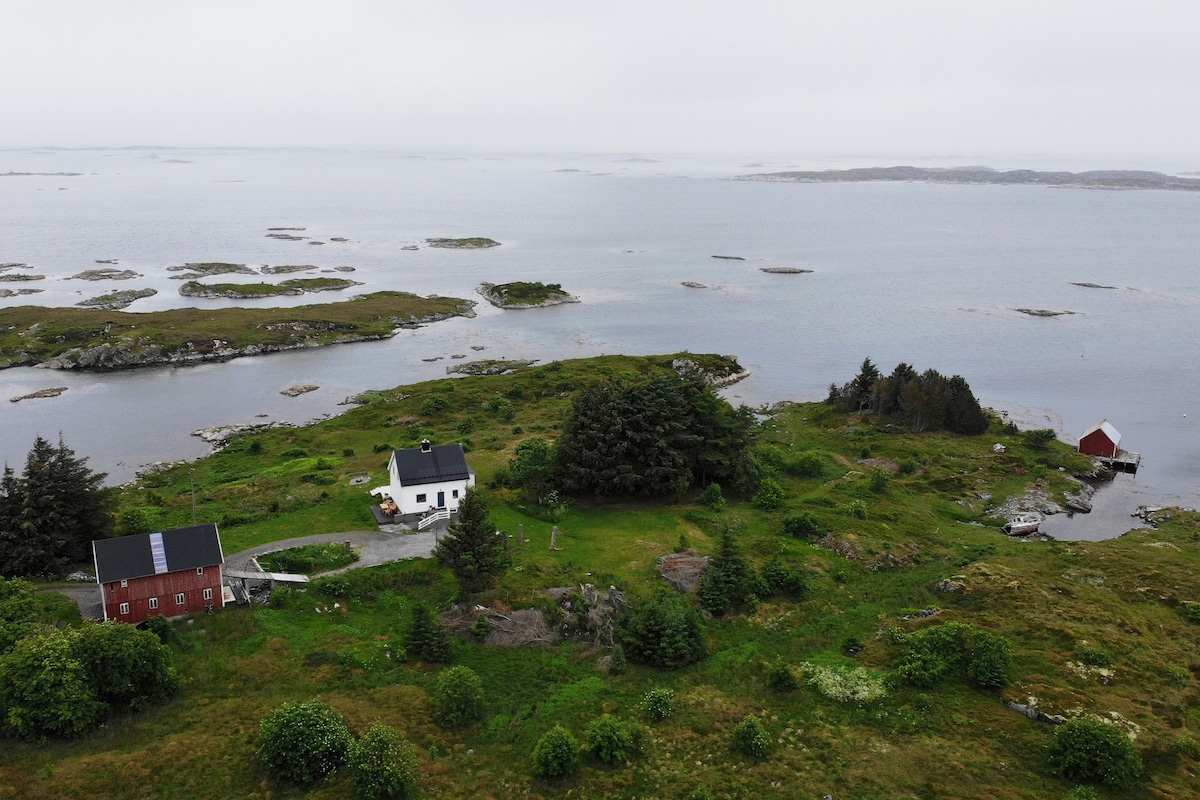 An aerial view captures a serene landscape featuring gently rolling greenery and a coastline dotted with small islands. Two picturesque buildings are visible, including a white house and a red boathouse, surrounded by lush vegetation and a calm bay.