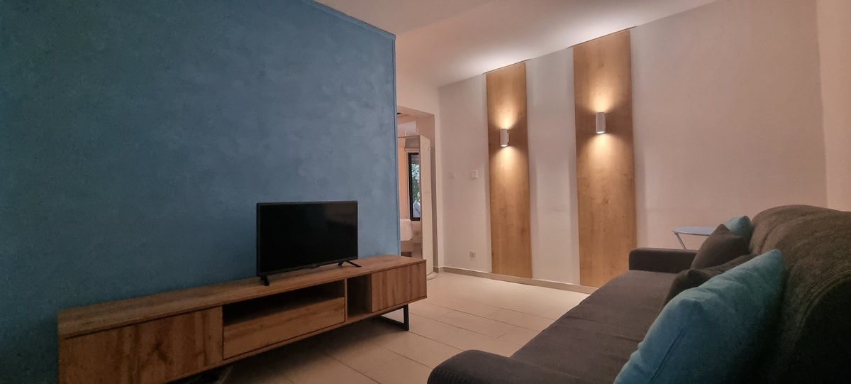 A cozy living area is featured, showcasing a flat-screen television on a wooden console. Two wall-mounted light fixtures illuminate the textured blue wall, while a comfortable sofa with decorative cushions provides seating. Neutral tones create a relaxed atmosphere in the space.