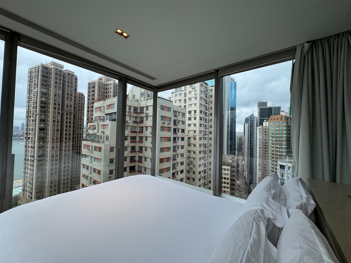 A spacious room is presented with floor-to-ceiling windows that offer views of a cityscape filled with tall buildings. The bed is neatly made with white linens, and natural light filters softly through the curtains, creating a serene environment.