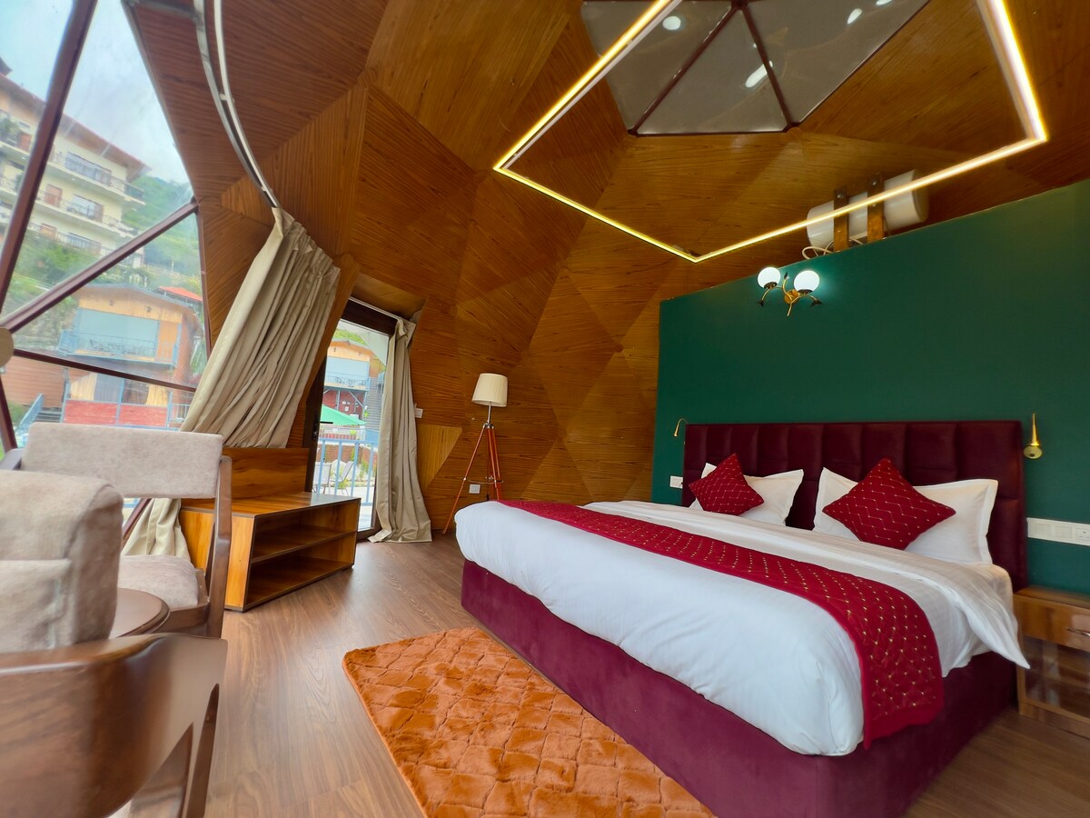 An inviting sleeping area features a spacious bed adorned with a deep red comforter and elegant accent pillows. A geometric wooden ceiling and large windows allow for an abundance of natural light. A cozy seating area is provided with a stylish chair and side table.