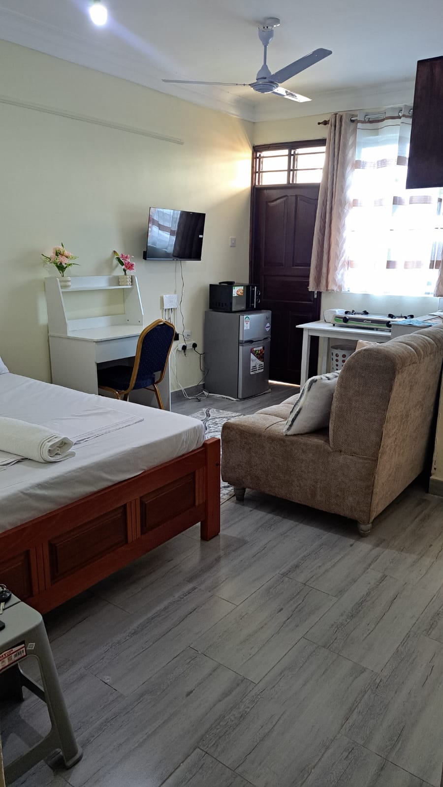A compact room features a spacious bed with neatly arranged linens. A television is mounted on the wall opposite the bed. A small desk and chair are positioned nearby, with a refrigerator situated next to a comfortable armchair. Natural light enters through a partially draped window.