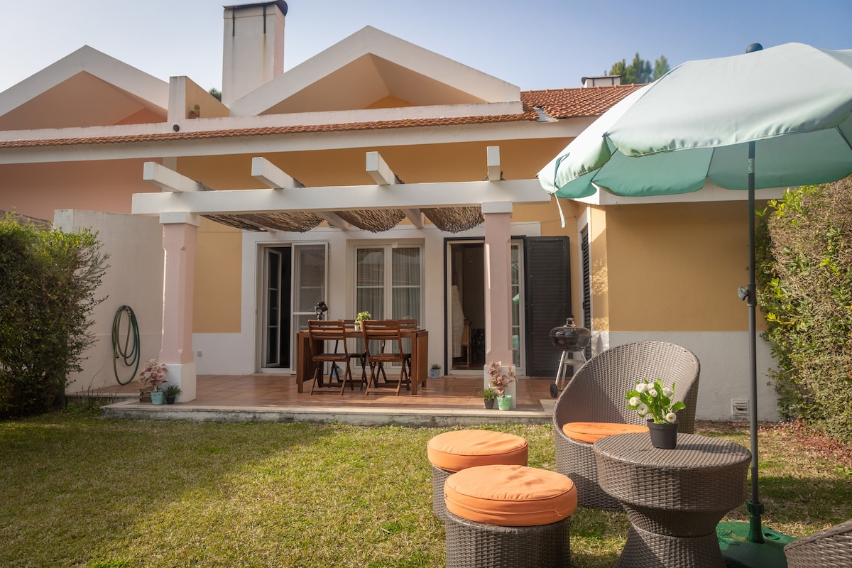 The image shows a charming outdoor area featuring a patio with a dining table and chairs beneath a pergola. A green umbrella provides shade over a seating arrangement with circular stools. Lush greenery surrounds the space, creating a serene atmosphere.