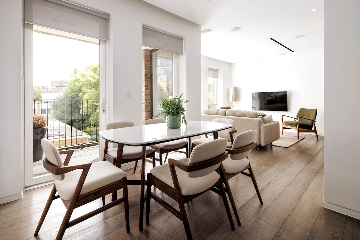 A modern dining area features a sleek white table surrounded by six cushioned chairs. A comfortable seating area with a sofa and armchair is positioned against a light wall. Large glass doors provide natural light and access to an outdoor balcony overlooking greenery.