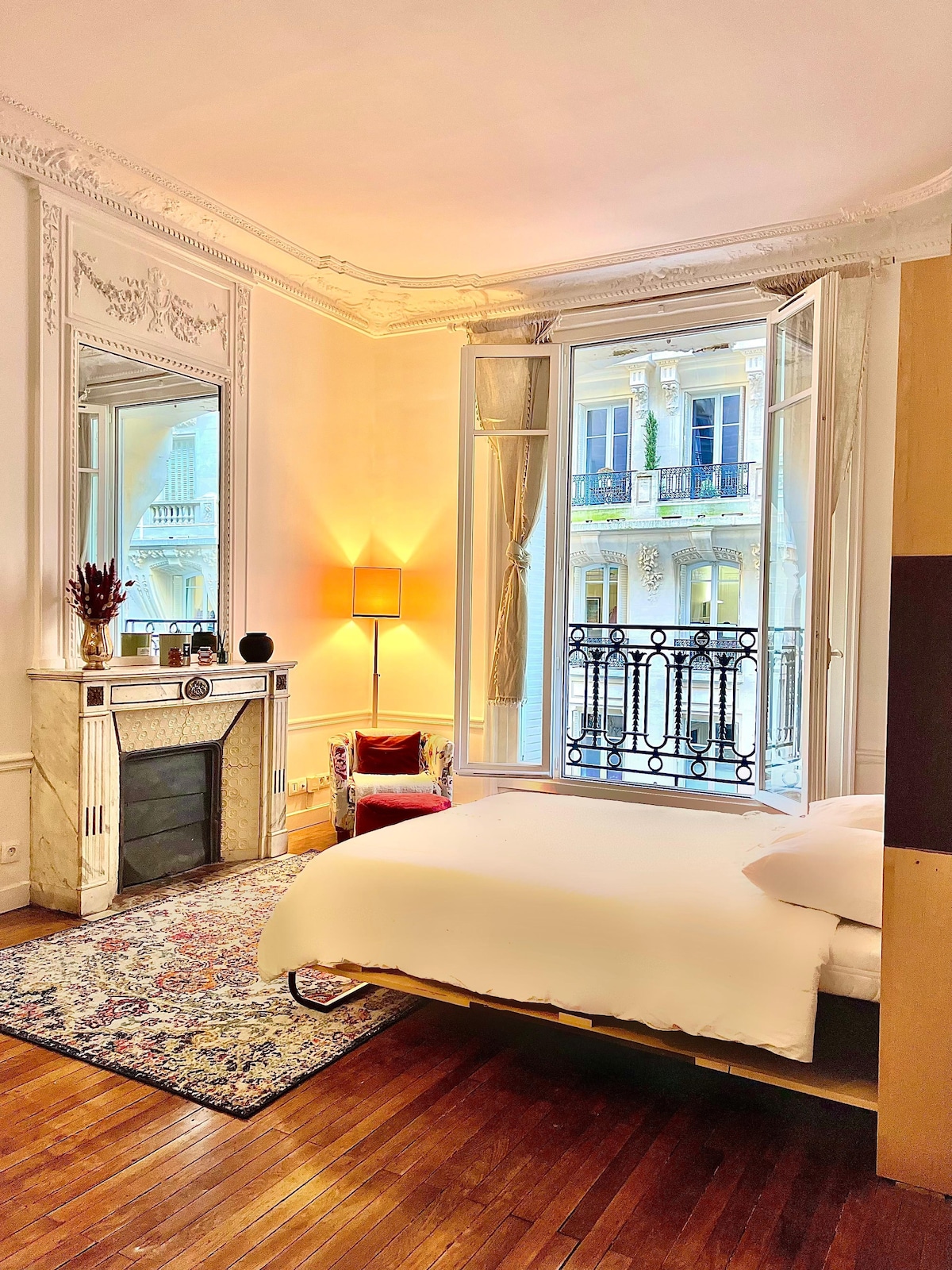 The studio features a cozy sleeping area, highlighting a Murphy bed with fresh linens. A decorative fireplace is positioned beside a full-length mirror. Light floods in through large windows with elegant drapes, revealing a view of a balcony and outdoor greenery.