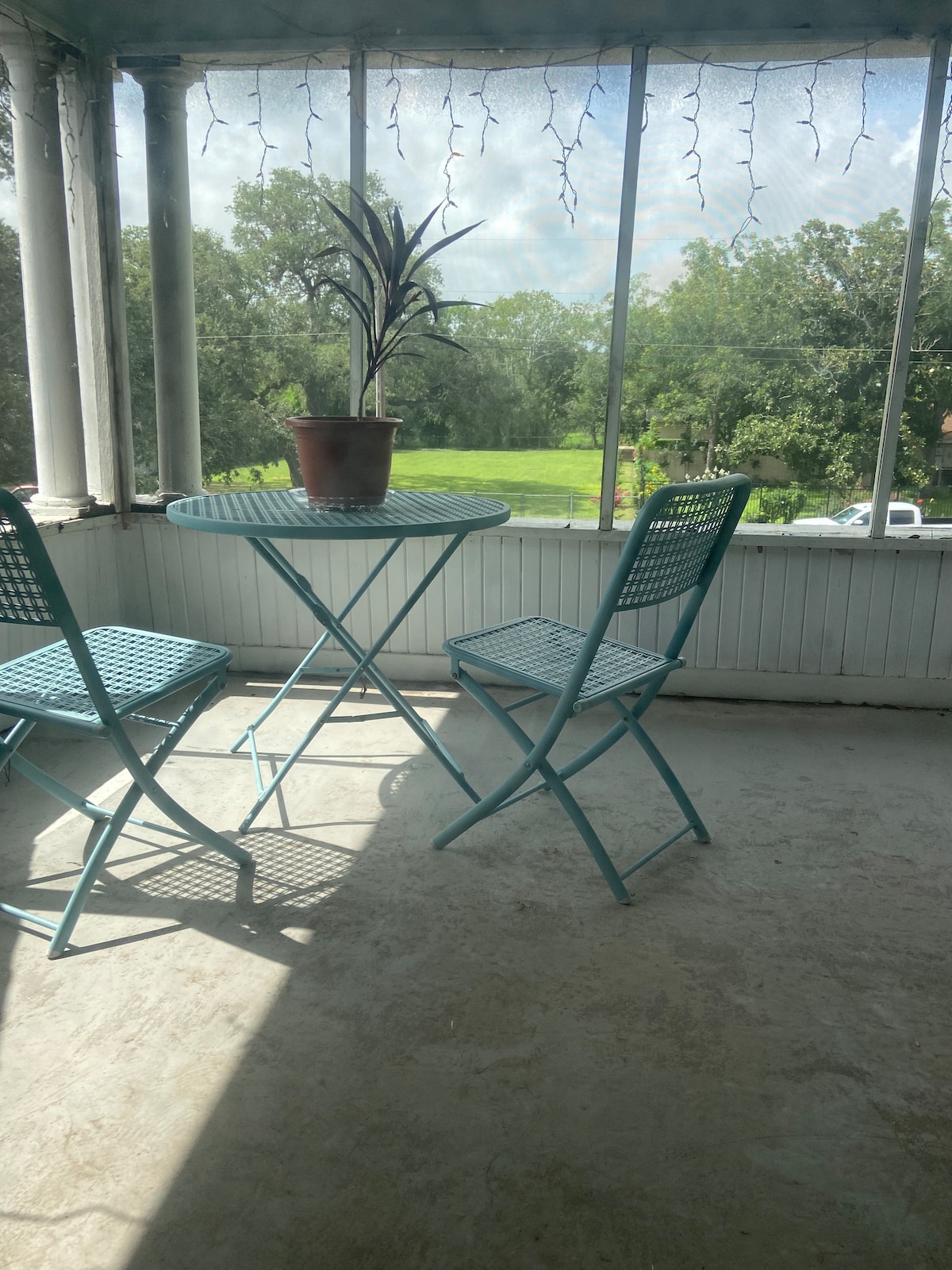 A sunny private patio is depicted with a round table surrounded by two blue metal chairs. A potted plant sits centrally on the table. Natural light filters through the mesh screens, illuminating the concrete flooring and green scenery visible beyond the patio.