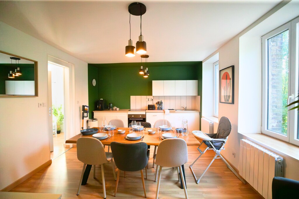 A modern dining area is featured with a large wooden table set with dinnerware. Soft lighting is provided by pendant lamps above. The kitchen, visible in the background, is equipped with sleek white cabinets and appliances. Large windows allow natural light to fill the space.