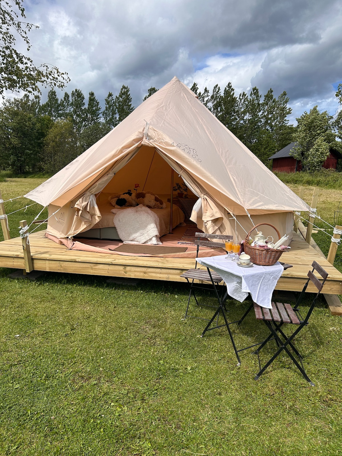 A spacious glamping tent is set on a wooden deck, featuring a neatly made bed inside. Outside, a small table and two foldable chairs are arranged, complementing a cozy outdoor setting. Lush greenery surrounds the tent, enhancing the natural ambiance.