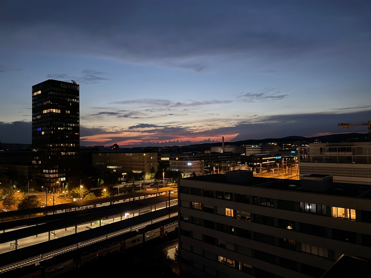 An evening skyline is depicted, showcasing a blend of urban structures against a gradient sunset. Streetlights illuminate the surroundings, and a train station is visible below, contributing to the city's vibrant atmosphere. The horizon features soft hues of orange and blue.