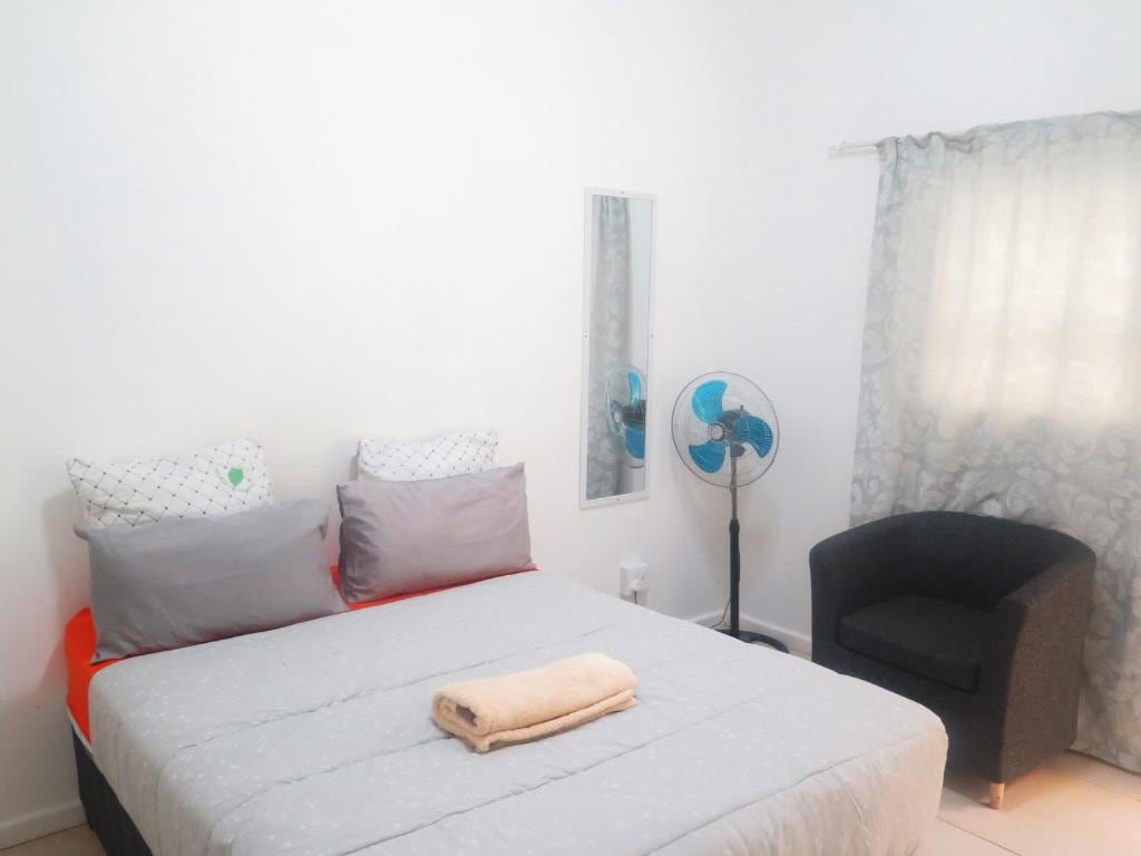 A comfortable bedroom is featured with a spacious bed covered in light linens. Two decorative pillows rest at the head of the bed. A standing fan is positioned nearby, and a mirror reflects the soft light coming through the partially draped window. A cozy chair is placed in the corner.