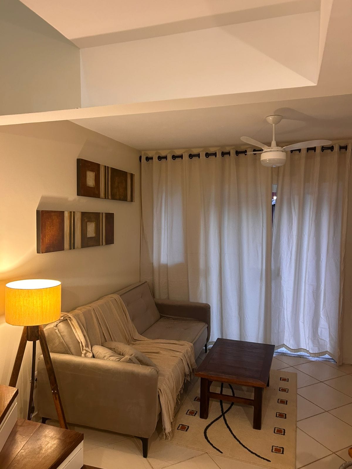 A cozy living area is presented with a neutral sofa draped in a light throw. A wooden coffee table is positioned in front, while a floor lamp emits soft light. Light-colored curtains frame a window, allowing natural light to fill the space.