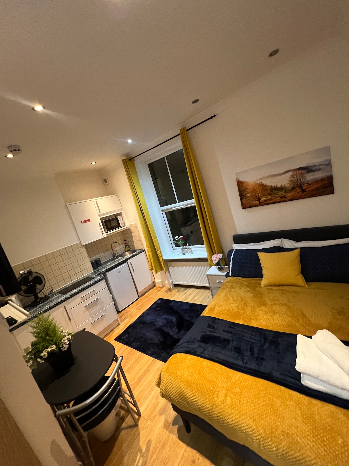A modern studio layout features a spacious bed with a vibrant yellow and navy bedding set. A compact kitchenette is visible with a microwave and sink. A small dining table with two chairs is positioned nearby. Light enters through a large window adorned with bright yellow curtains, enhancing the cozy atmosphere.