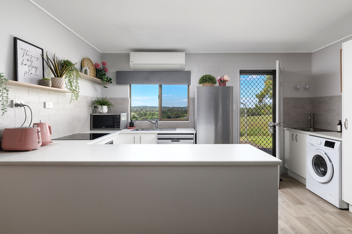 A modern kitchen features a clean layout with a central island and a mix of appliances including a microwave, kettle, and washer. Natural light floods the space through a large window, which offers expansive views of the outdoors. A door leads to the exterior.