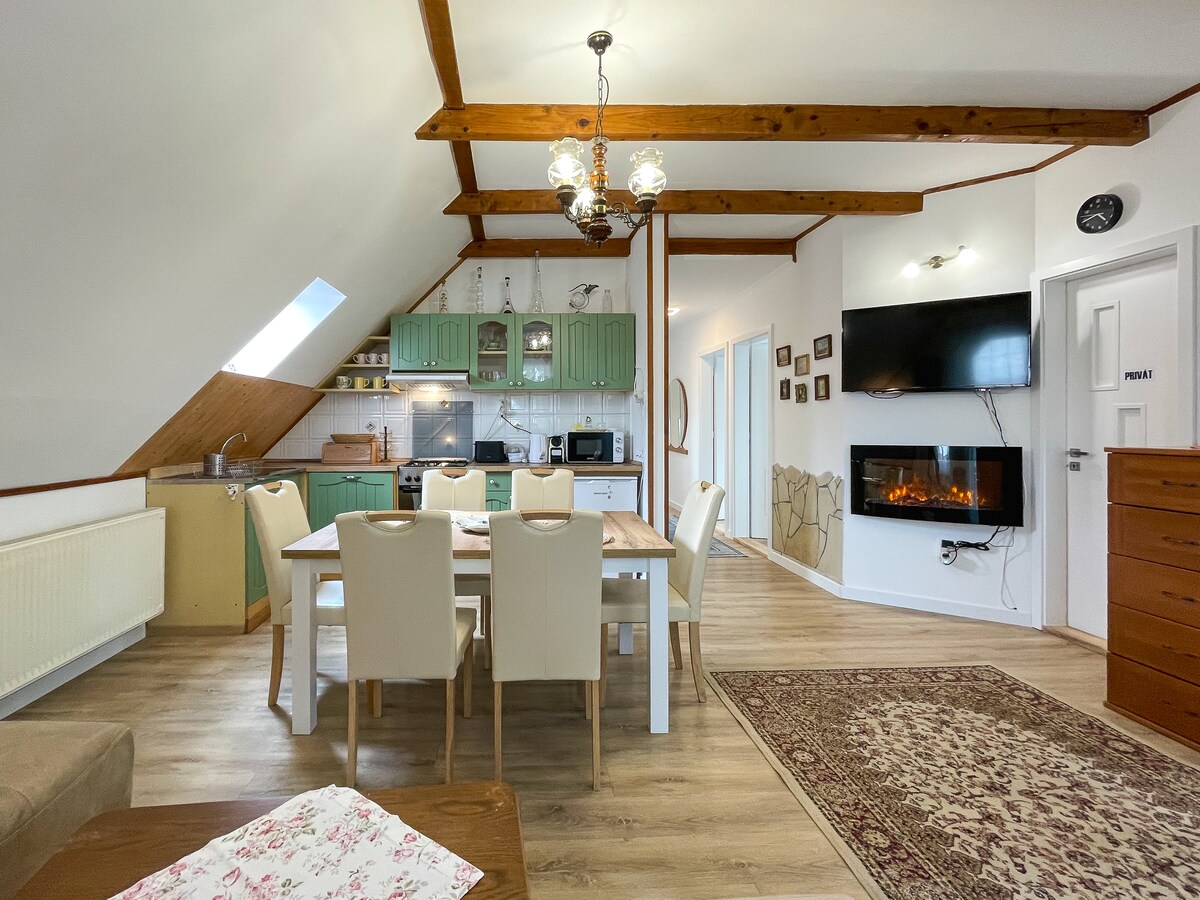 A spacious living area features wooden beams along the ceiling, a large dining table with white chairs, and a kitchen area with green cabinetry. A mounted flat-screen TV is positioned on the wall opposite the table, and a cozy electric fireplace adds warmth to the space.