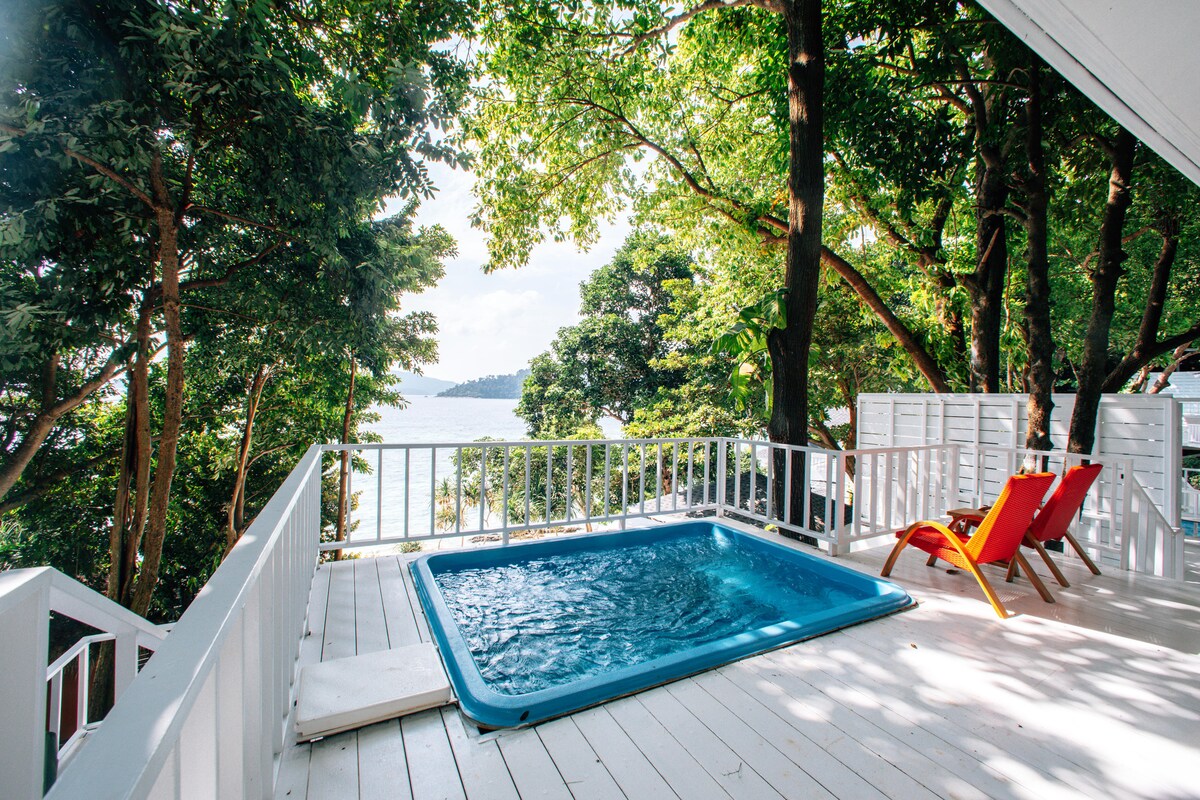 A private pool surrounded by a wooden deck is visible on a bright, sunny day. Two colorful chairs are positioned nearby, overlooking a lush area of trees and a view of the water beyond.