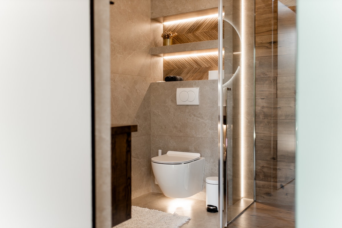 A modern bathroom features neutral-toned walls with soft lighting. A sleek white toilet is positioned against textured tiles, while a plush rug lies on the floor. The glass enclosure surrounding a shower is visible, adding to the contemporary design.