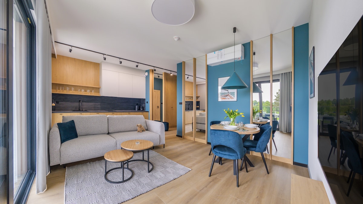 A modern living area showcases a light-colored sofa paired with two blue velvet chairs and a round wooden table. Natural light enters through large glass doors, illuminating the space against a backdrop of soft, neutral tones and a vibrant blue accent wall.