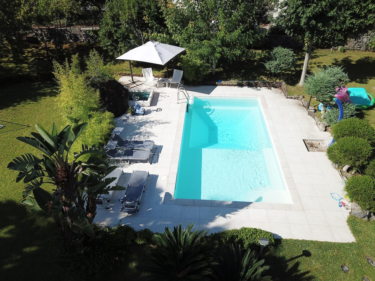 A private swimming pool is surrounded by a well-maintained garden, featuring lush greenery and a variety of plants. Sun loungers are placed around the pool, and a large umbrella provides shade. The pool's clear water reflects natural light, enhancing the serene outdoor setting.