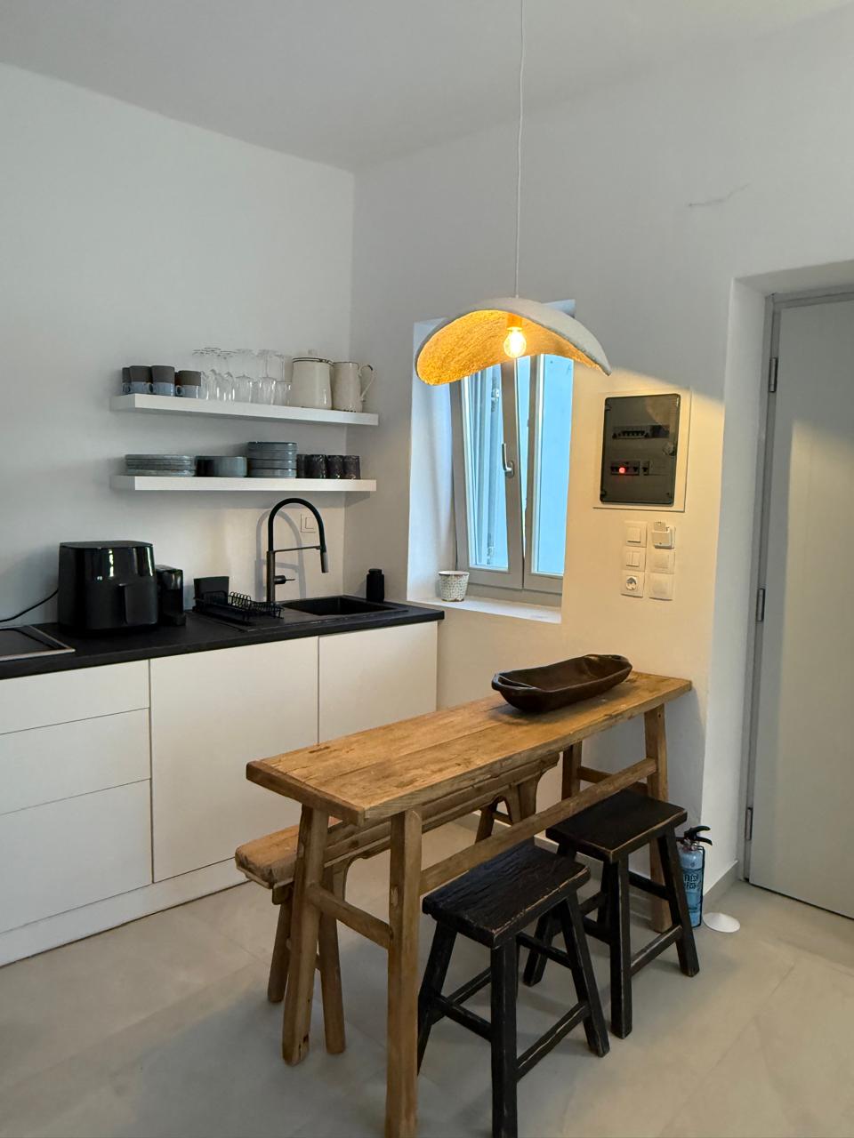 The kitchen features a sleek design with white cabinetry and a black countertop. A wooden dining table with three stools is positioned against the wall. Natural light fills the space through a window, enhancing the functional layout with modern appliances and minimalistic decorative elements.
