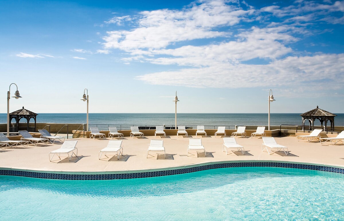An inviting pool area overlooks the ocean, featuring a spacious pool surrounded by lounge chairs. Gazebos provide shaded spots, and bright blue skies meet the calm waters in the background, creating a relaxing atmosphere for guests to enjoy.