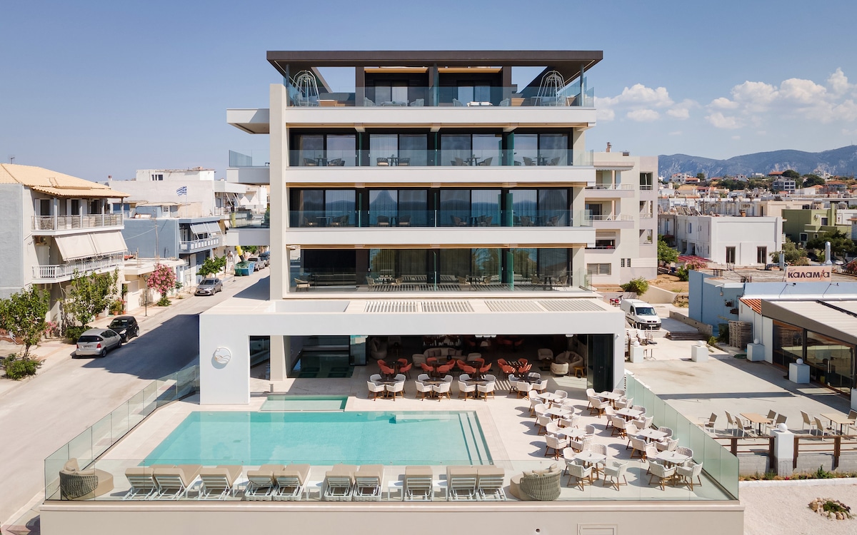 The Avant Blue Boutique Hotel is displayed from an aerial perspective, showcasing its modern architecture and multiple floors. An outdoor pool is featured at the base, surrounded by sunbeds and seating. The surrounding area reflects a vibrant street, adding to the overall setting.