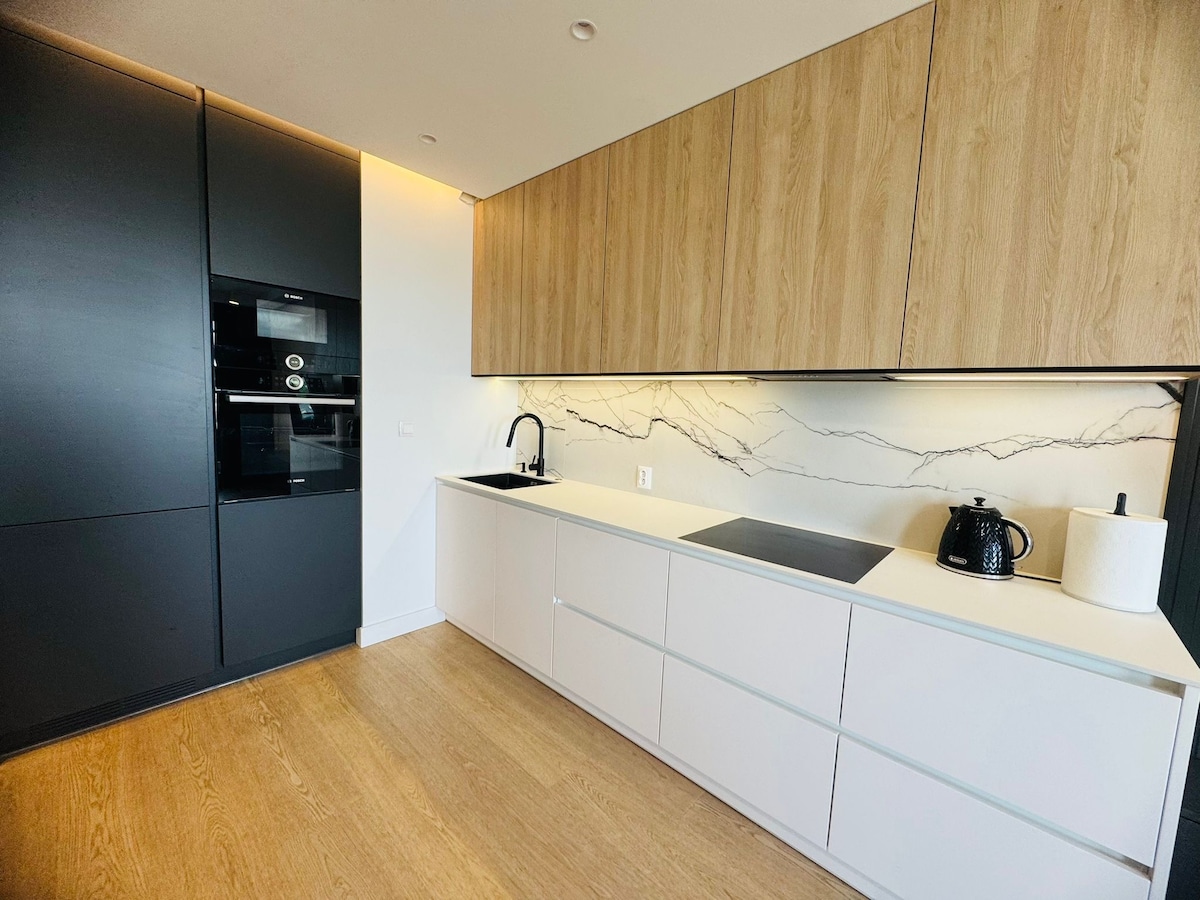 A modern kitchen is equipped with sleek cabinetry and a minimalist design. The space includes a black oven, a spacious countertop, a stylish kettle, and a white canister. The marble backsplash adds a sophisticated touch, while wooden accents create warmth.