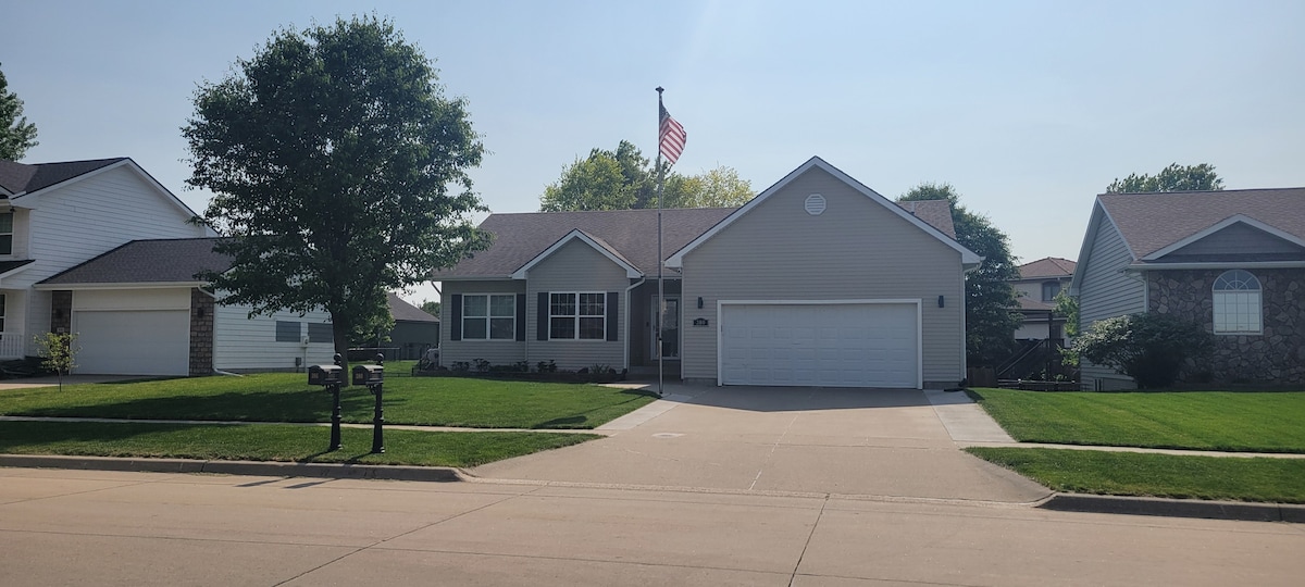 A single-story home is displayed with a neatly landscaped front yard featuring a well-maintained lawn. An American flag stands proudly on a flagpole. A two-car garage is visible, alongside a wide driveway. The surrounding area includes similar residential homes, creating a friendly neighborhood atmosphere.