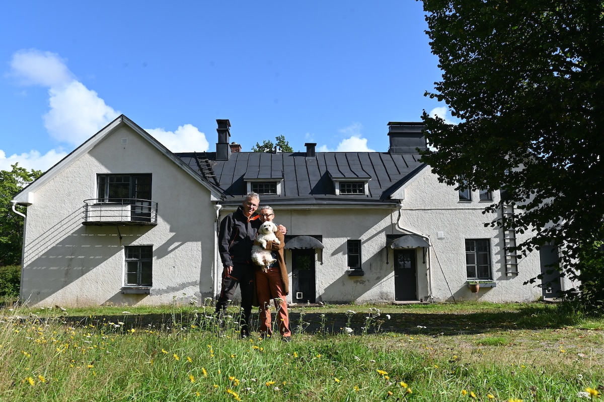 A two-story stone building with a grey roof and white walls is showcased against a clear blue sky. The structure features multiple windows and a balcony. A grassy area laden with wildflowers surrounds the house, creating a natural setting for visitors.