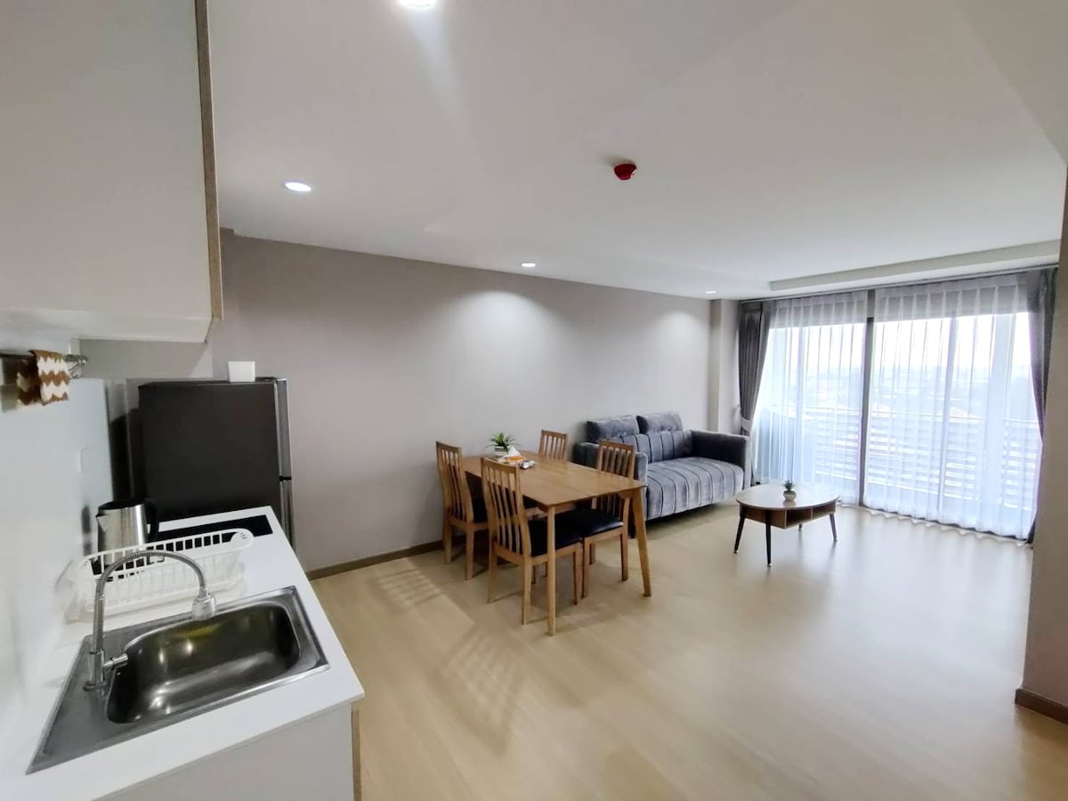 An open living space features a kitchenette with a sink and dining table surrounded by light wood chairs. A comfortable gray sofa is positioned near large windows, allowing natural light to brighten the area. The overall atmosphere is calm and inviting.