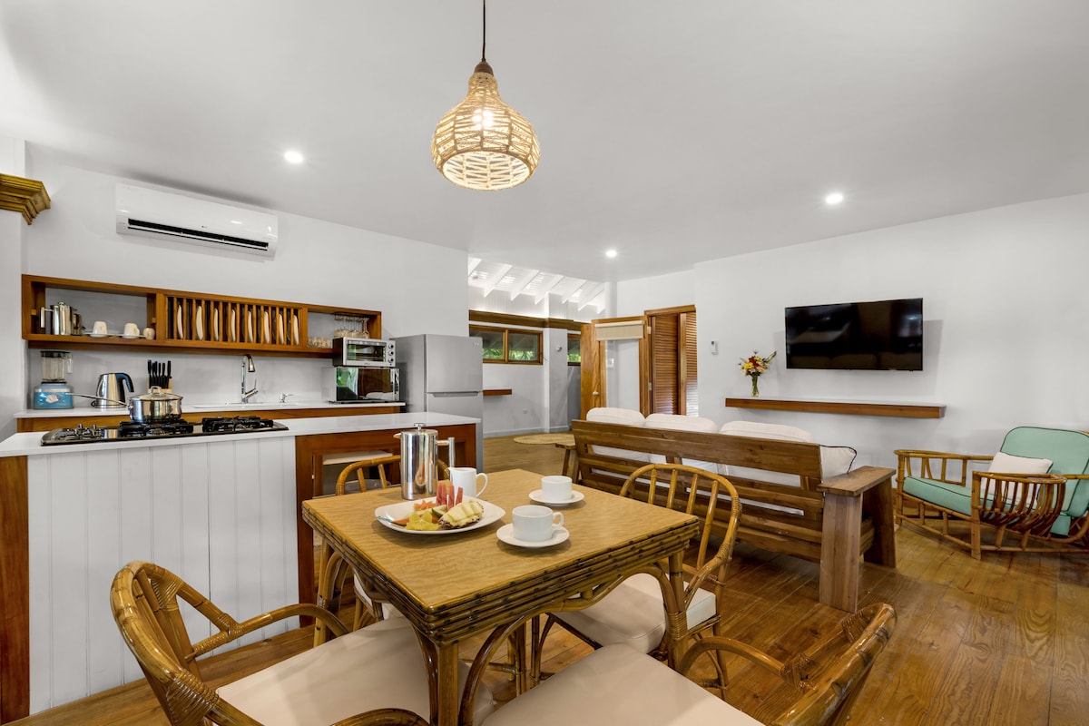 An open and airy living space features a wooden dining table set for two, accompanied by comfortable chairs. A well-equipped kitchen is visible in the background, with modern appliances and wooden shelving. A cozy seating area includes a television and bright, inviting decor.