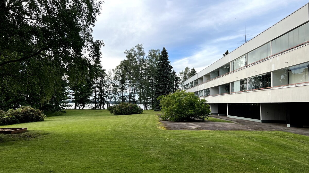 A spacious lawn is surrounded by trees, providing a serene outdoor area. A modern building with multiple windows is visible on the right, with a smooth pathway in front. The scene captures a peaceful environment near the water's edge, enhancing the natural landscape.