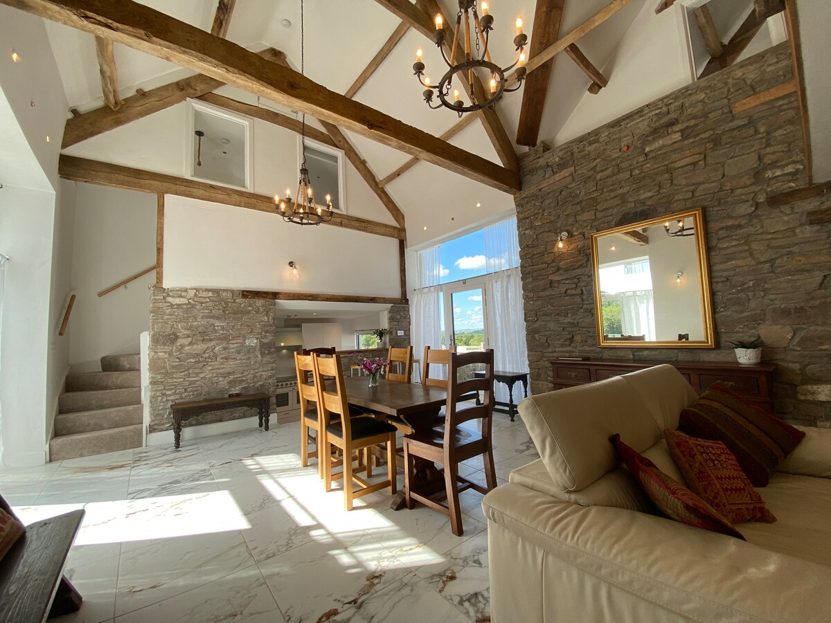 A spacious living area showcases a blend of natural stone walls and wooden beams. A large dining table is surrounded by several chairs, and light streams in from large windows. A cozy seating arrangement is positioned nearby, enhancing the inviting ambiance.