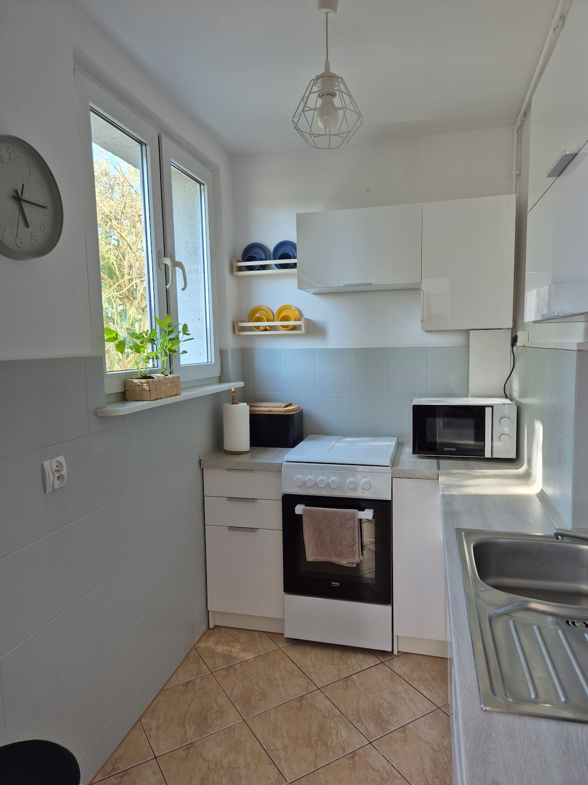 A well-equipped kitchen features a stove with an oven, a microwave, and a sink set into light grey cabinetry. Natural light filters through a small window, accentuating the clean lines of the space. Shelves hold colorful dishes, complemented by neutral-toned decor.