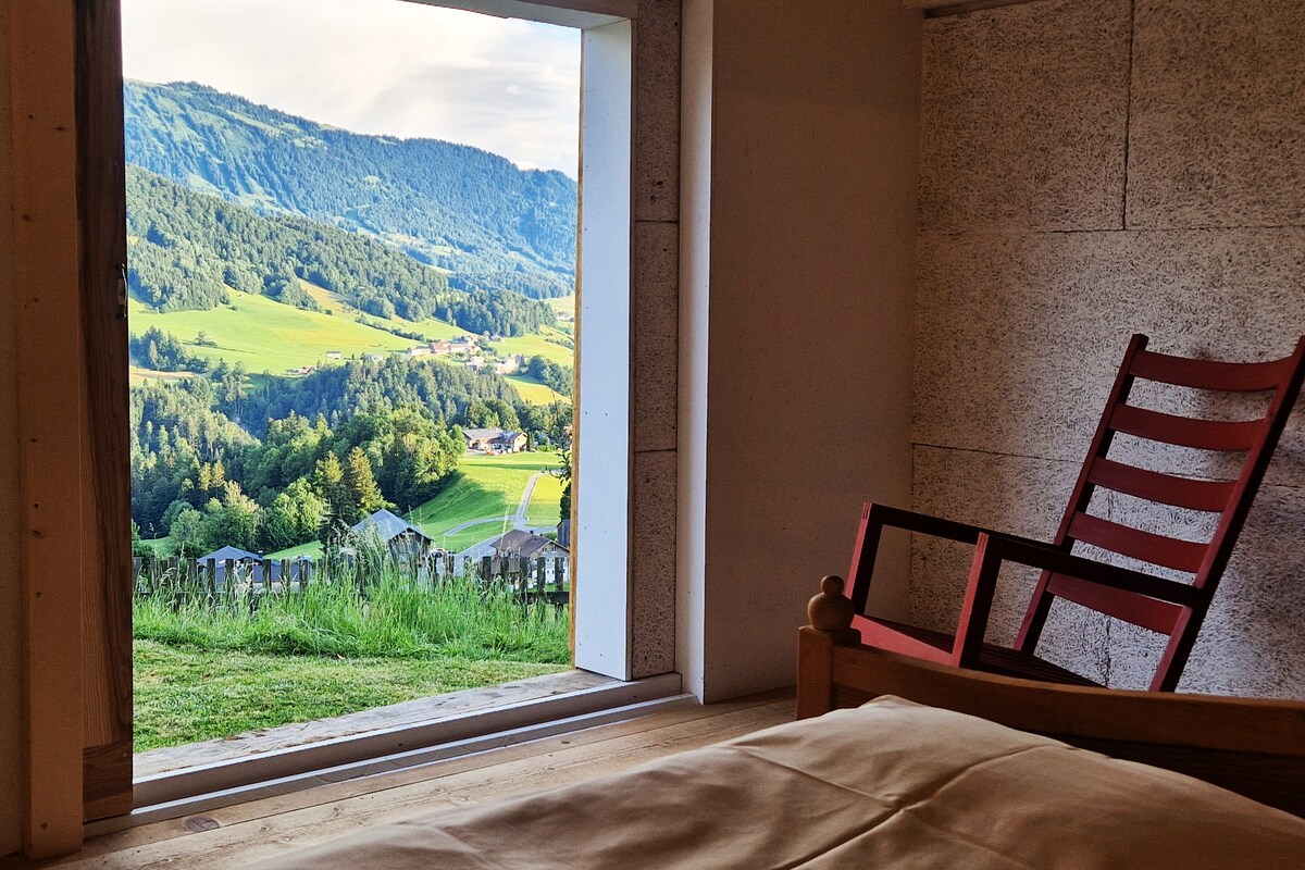 A spacious bedroom is highlighted by a large window framing a stunning view of rolling hills and lush greenery. A simple wooden bed is positioned in the foreground, while a red chair rests nearby, providing a cozy spot to enjoy the scenery.