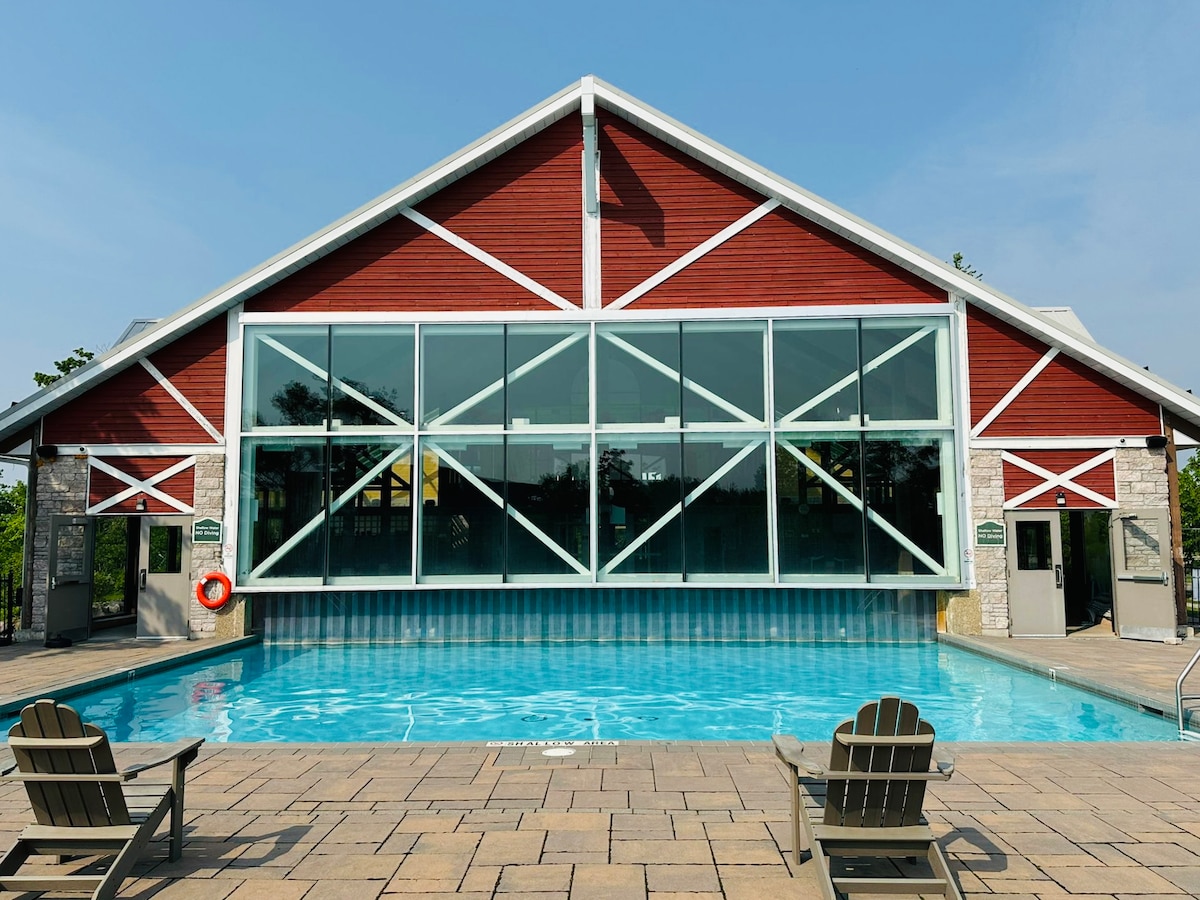 A spacious pool area is framed by a modern red and white building featuring large glass windows. The inviting turquoise water reflects the sky. Two wooden lounge chairs are positioned alongside the pool, creating an ideal spot for relaxation.