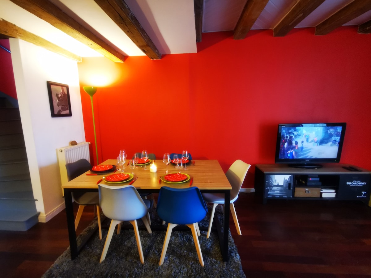 A dining area features a wooden table set for a meal, with colorful plates and glassware arranged on top. A cozy seating arrangement includes four chairs in various colors. The wall is painted a vibrant red, complemented by warm wood beams above, and a television is visible in the corner.