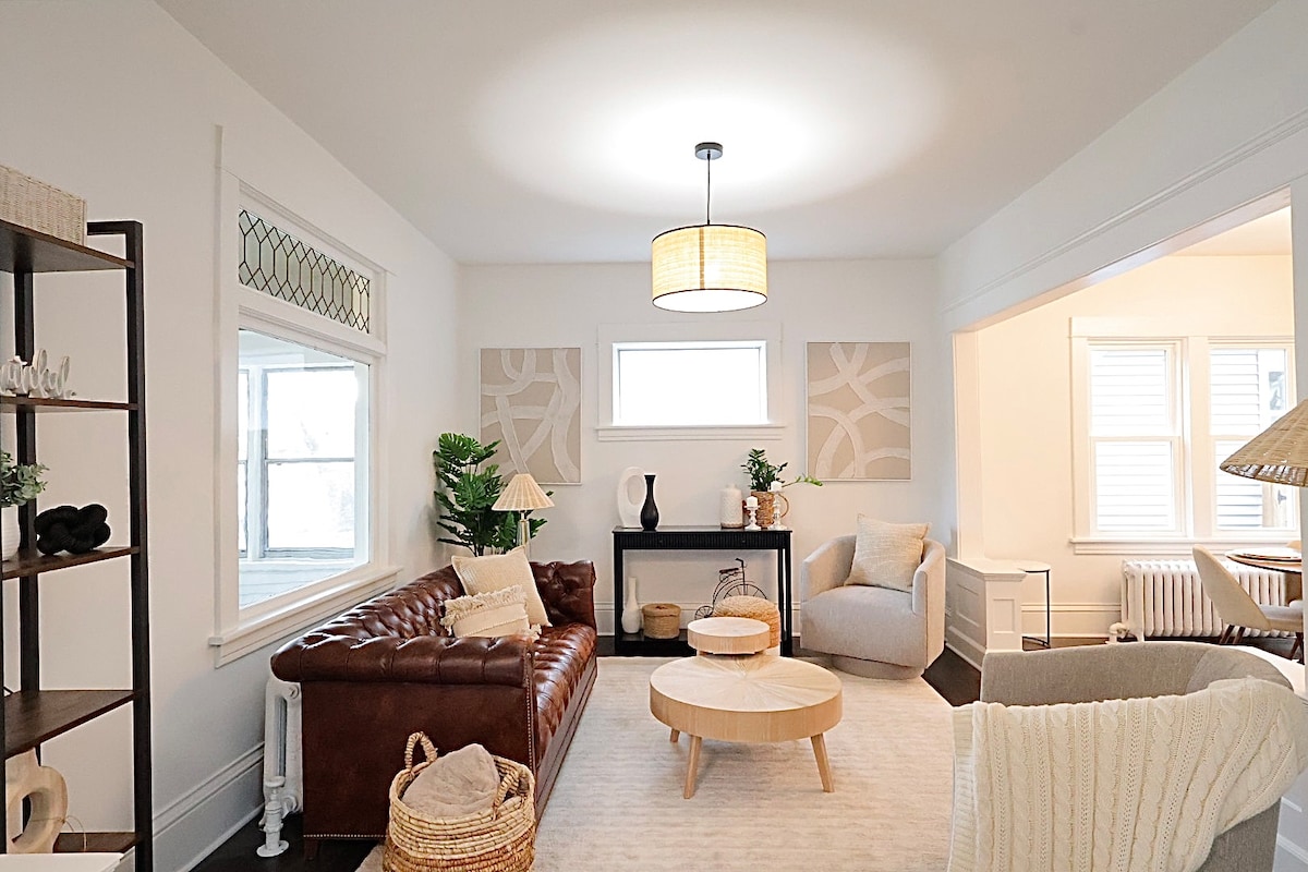 A bright living room features a combination of modern and classic elements. A leather sofa and two light-colored chairs are arranged around a round coffee table. Natural light fills the room through two windows, complemented by neutral decor and houseplants.