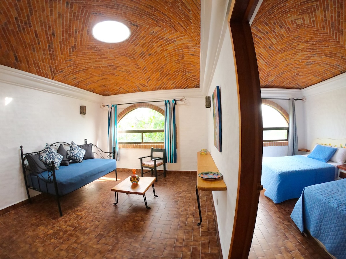A bright and airy room features high, arched brick ceilings with natural light streaming through large windows. Two beds are present, along with a daybed and a wooden coffee table. Colorful cushions add a touch of vibrancy, while curtains frame the windows.
